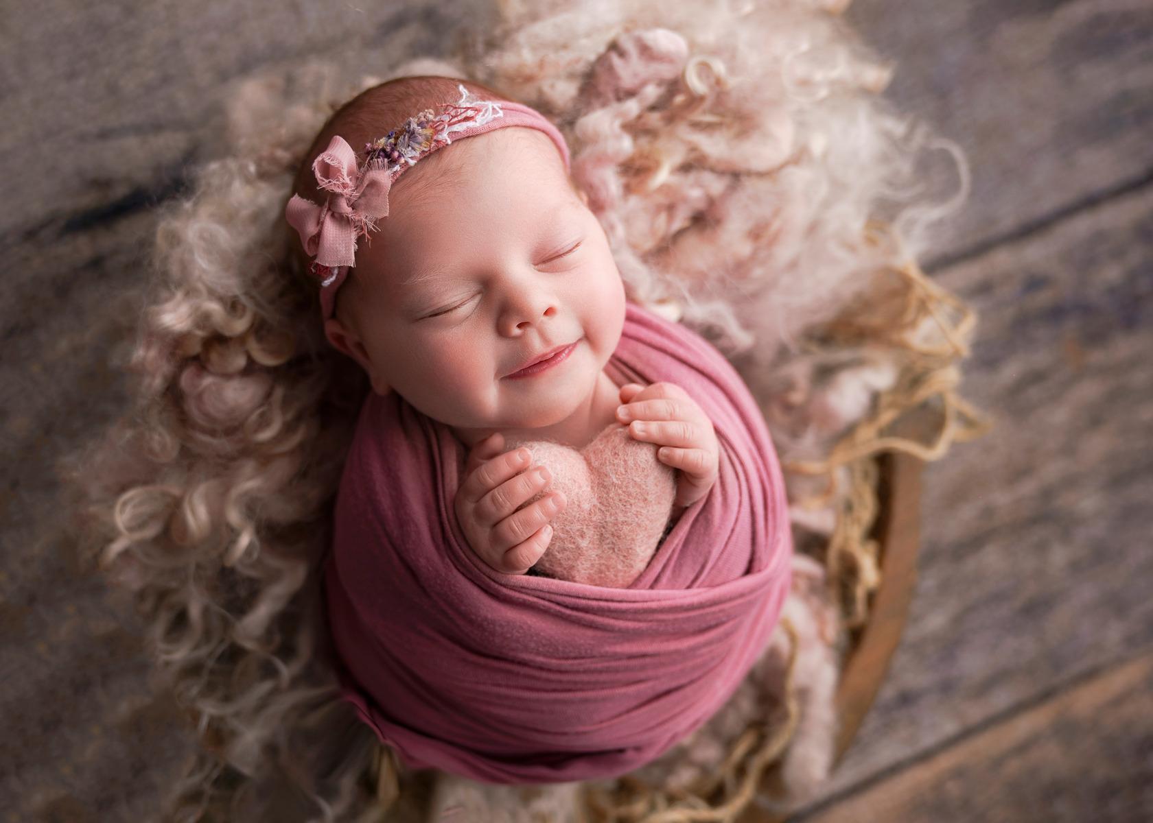 "Adorable baby girl wrapped in pink holding a heart prop, wearing a cute pink headband, photographed by Ceire Rhein, Dublin's newborn photographer."
